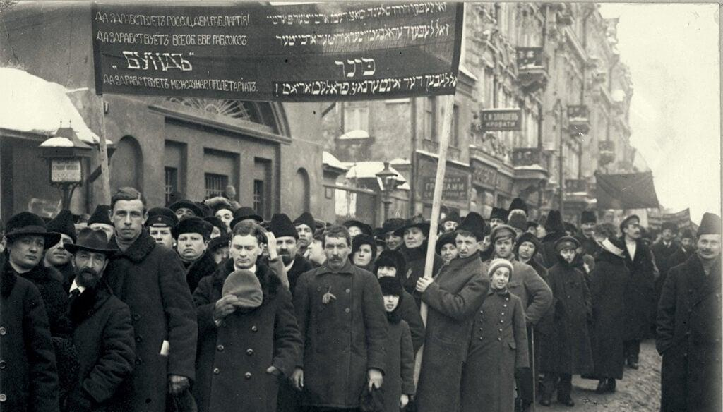 A black-and-white photograph of a large group of people of mixed ages ranging from children to a man who appears to be in his 50s or 60s, wearing mostly black and dark grey coats and black hats of various shapes and sizes, walking in a large group through a street, as in a demonstration. They carry signs and banners, the most visible made of dark fabric hoisted between two tall wooden poles. It contains lettering in white Yiddish and Cyrillic script. A series of building facades stands behind and to the left of the crowd. Bright white snow is visible on some of the peaks of the buildings and on some lampposts.